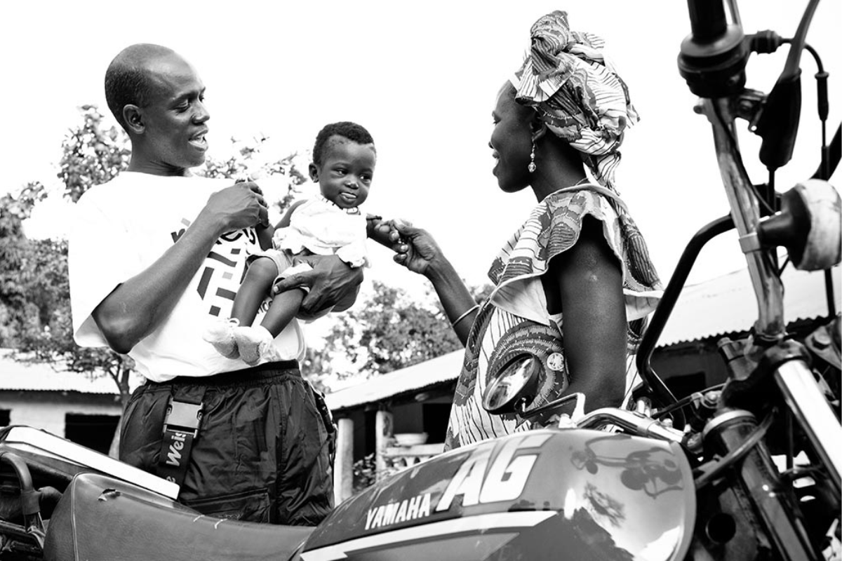 Black and white image of a smiling man holding a baby while a woman in patterned traditional attire reaches out to the baby. A Yamaha AG motorcycle is partially visible in the foreground.